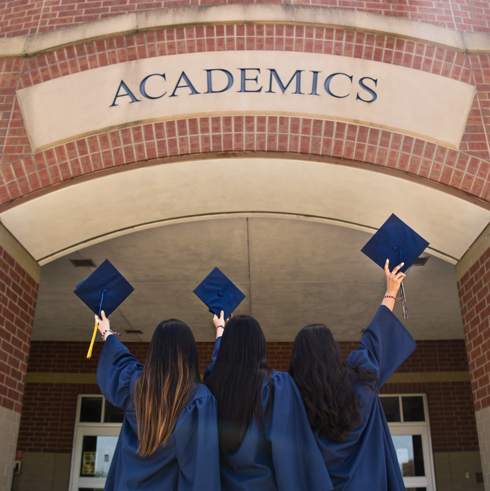 Rear view of three students in graduation gowns holding their hats up to a sign saying "Academics"