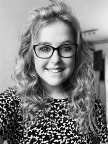 Black and white photo of a young white woman with pale curly hair and large glasses smiling into the camera
