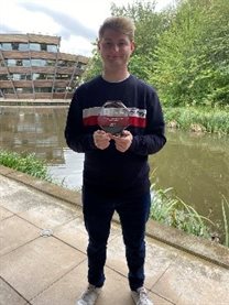 Placement Awards Prize Winner Shaun Hoskins, stood by the lake on Jubilee Campus holding his award. Jack is wearing dark trousers and a dark long sleeve top with a white and red horizontal stripe across the chest..