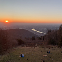 A sunset view over a valley, a river and city can be seen in the distance and couples picnicking in the foreground