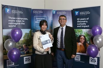 Winner Chi (Rosy) Nguyen holding a certificate with Dr Amrit Judge standing in front of NUBS banners with balloons attached