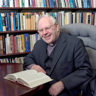 Anthony Thiselton sits smiling at a desk in front of a bookshelf with an open book before him on a table