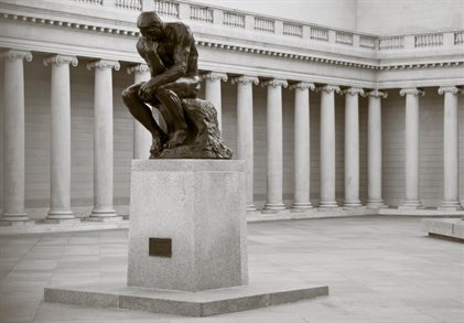 he Thinker statue, a bronze statue of a man with his elbow on his knee and chin resting on his fist outside a building