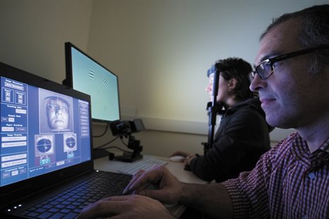 A researcher sits at a laptop monitoring an eye-tracking experiment while the participant is undergoing a test