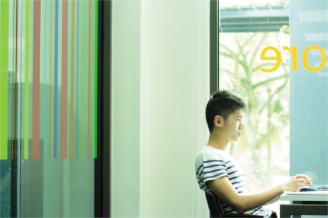An asian student sits at a desk with his laptop, in front of a large window