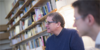 Two bespectacled men sit in front of a wall of neatly shelved books taking part in a discussion