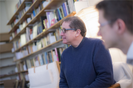 Two bespectacled men sit in front of a wall of neatly shelved books taking part in a discussion