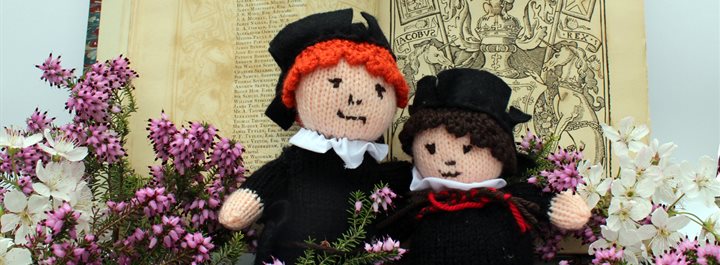 Photo of two knitted soft toys (modelled on Douglas and Boece) leaning against an open book surrounded by sprigs of heather. Gavin Douglas and Hector Boece were major figures of Scottish humanism.