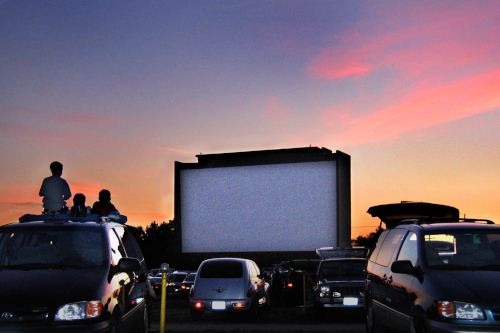 People sitting on top of cars to watch a film in an outdoor cinema during a sunset.
