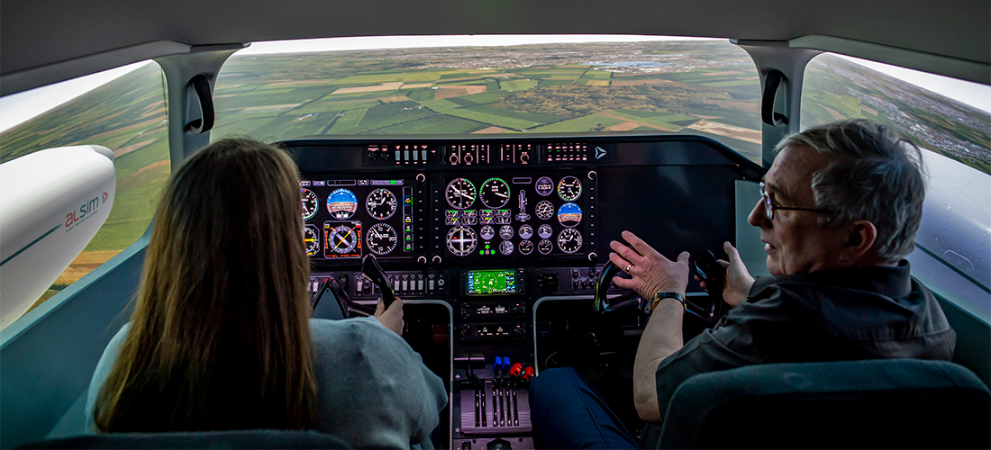 Student piloting the flight simulator, instructed by an academic, with a simulated landscape visible through the cockpit windscreen and windows