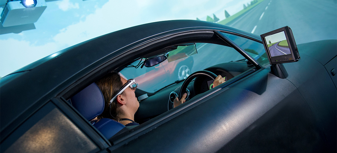 Student sat at the steering wheel of a car with a projector overhead and a simulated road and landscape visible around it