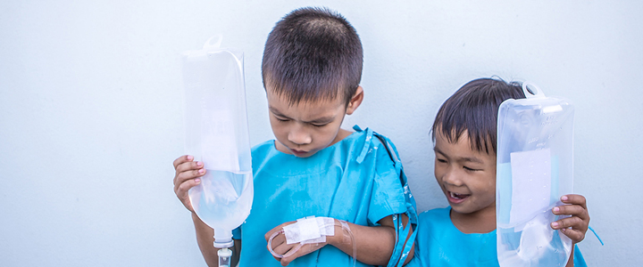 Two young children in hospital gowns holding up and looking at their IV drips