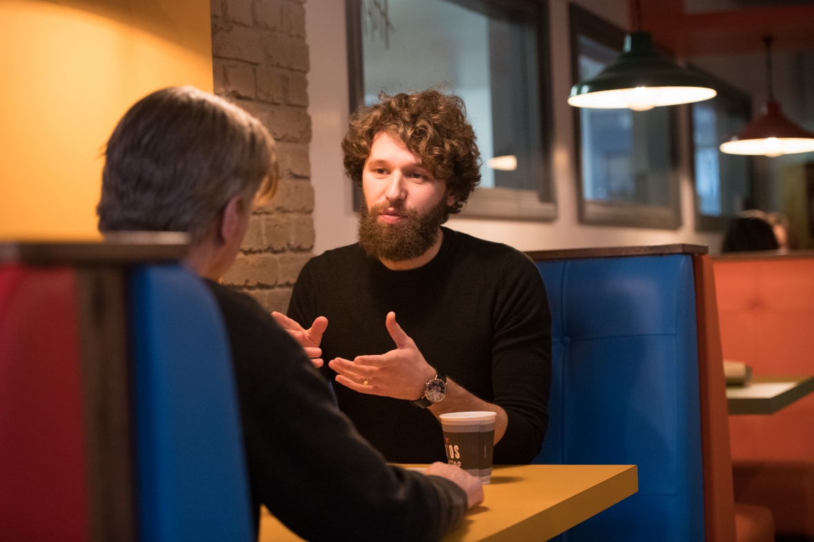 Male student with curly hair and a beard, talking to another person in a cafe