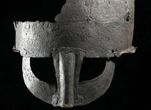 Photograph of a a rusted and partially missing ancient helmet with a plain curved helm, noseguard and covers around the eyes against a black background.