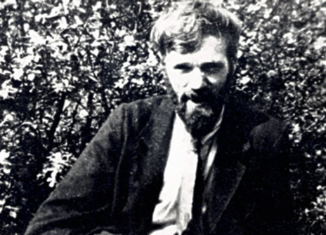 Black and white photograph of a white man with a beard seated in front of a hedge.