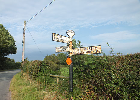 Photograph of an old metal signpost with white arms pointing to local towns in three directions. The signpost is located in the grass verge next to a country road with a hedgerow and blue skies behind.