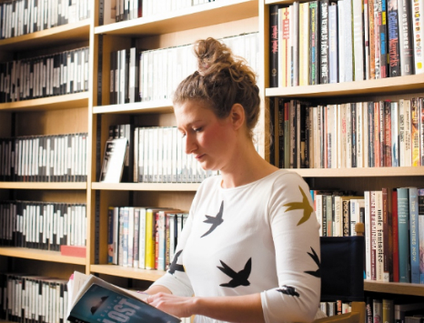 Photograph of a young white woman in profile wearing a white jumper with a large bookcase filling the wall behind her.