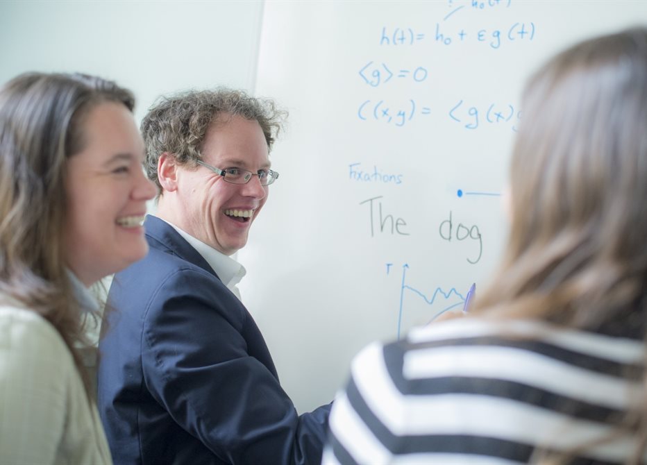 Photo of two white women and a man stood in front of a whiteboard with writing on it and laughing together.