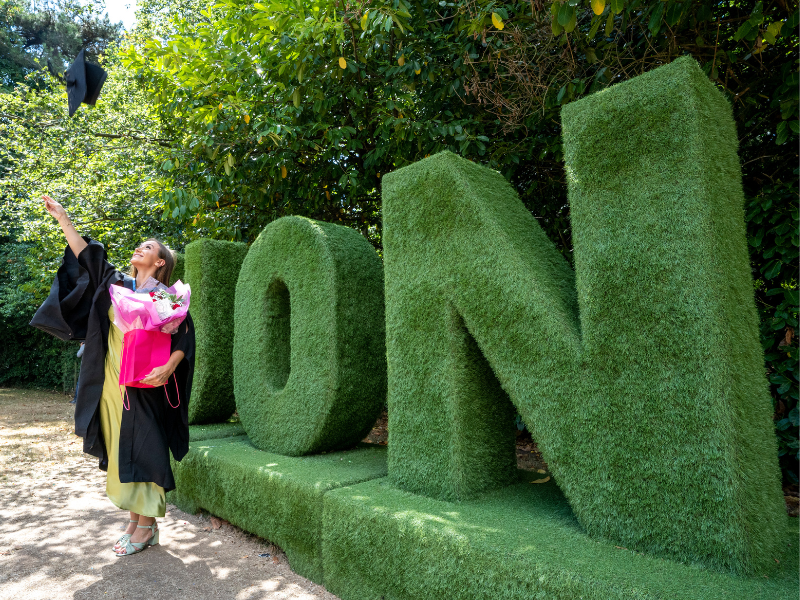 Photograph of a graduate in front of the 'UoN' grass lettering