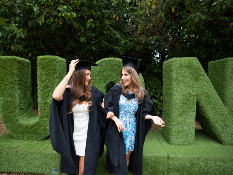 Photograph of two graduates smiling in front of the 'UoN' grass lettering
