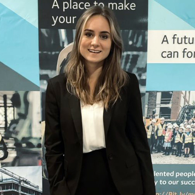 Emma Sharman wearing a black suit and smiling in front of a banner