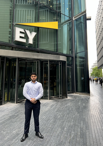 Neyaz Zaman in a smart shirt and grey trousers standing in front of a large glass building with EY logo