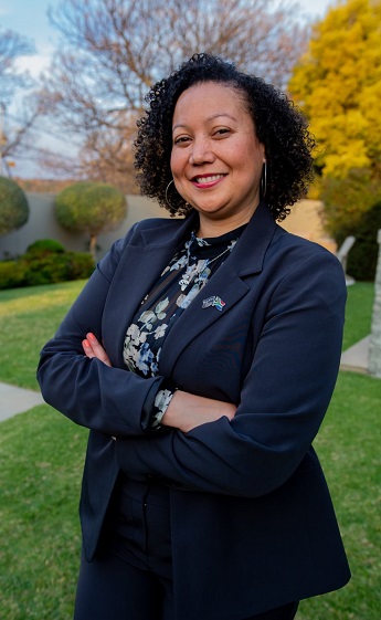 Photo of MBA graduate Mecia Peterson. Mecia is stood outside with trees visible in the background. Mecia is wearing a dark navy jacket and a floral top and is folding her arms across her body.