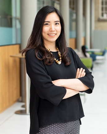 Image of Pai smiling. Wearing a black top and wooden bead necklace with arms folded.