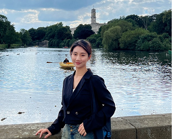 Chinese student Zixuan Xiao standing in front of the lake at university Park campus with the clock tower in the back. Wearing jeans and a dark blue jacket.