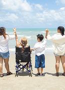 Four people on a tropical beach looking at the ocean. They are holding hands with their arms up in the air. The second person from the left is in a wheelchair.