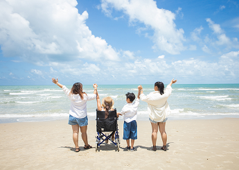 Four people on a tropical beach looking at the ocean. They are holding hands with their arms up in the air. The second person from the left is in a wheelchair.