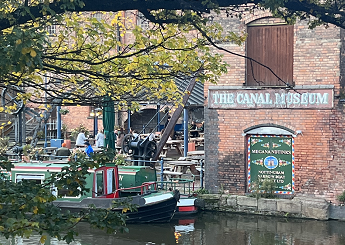 A canal-side cafe with canal boats in the foreground and tables in the background. The building displays the words Canal Museum.