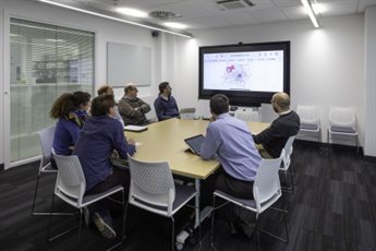 An executive board sit around a table in a meeting room and look at a presentation screen on a wall.