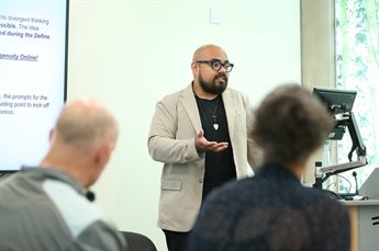 Jose Gonzalez Lopez teaching a programme. He stands next to a lectern with a slideshow to one side. Two delegates, one a white male and one a black female who are visible from behind, listen to him speak.