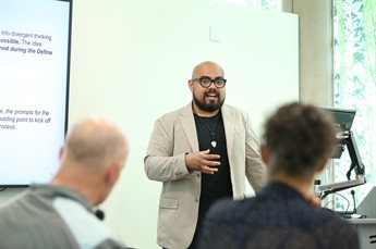 Jose Gonzalez Lopez teaching a programme. He stands next to a lectern with a slideshow to one side. Two delegates, one a white male and one a black female who are visible from behind, listen to him speak.