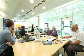 5 participants and a a member of the delivery team sit around tables with laptops during a training programme. Jose Gonzalez Lopes stands at the front of the room gesturing to a slideshow as he delivers the content.