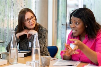 A female staff member listens to a black programme participant talk during a discussion session. They sit side by side at a table and the participant talks animatedly, leaning forwards slightly and using using emphatic hand gestures. The staff member list