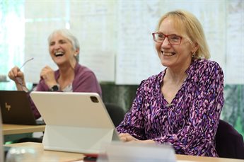 Two older women sit side by side during a programme. They both have laptops on the table in front of them and they are laughing.