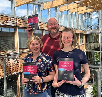 ICCSR members Lee Matthews, Lara Bianchi, and Claire Ingram holding a copy of the Concise Encyclopaedia of Corporate Social Responsibility standing in the Business School North atrium in a sunny day