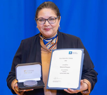 Professor Meryem Duygun holding Vice-Chancellor’s Medal 2024 and a certificate standing in front of a blue curtain.