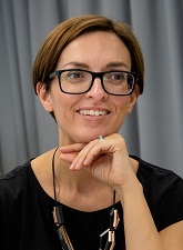 Photo of Sarah King. Sarh is wearing glasses, a black short sleeve top and a necklace. She is smiling and resting her chin on her hand. cin on her