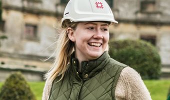 Photo of a smiling young white woman with blonde hair wearing a hard hat. There are old walls and a green lawn in the background.