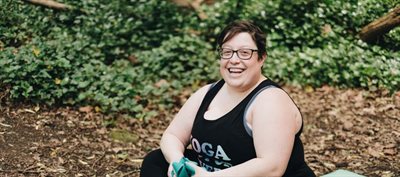 Photograph of a smiling white woman with glasses, short hair and wearing a yoga tank top seated outside in a wood
