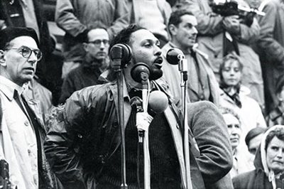 A black and white photograph of an activist speaking to a crowd in front of three microphones.