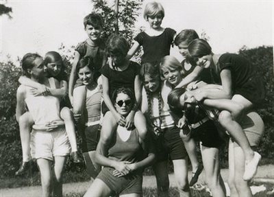 Black and white photograph of a family gathered for a photo. Younger children are piggy backing, or on the shoulders of taller children and adults. They&amp;#39;re wearing summer shorts in a garden.