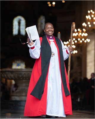 Bishop Rose of Dover, smiling in the foreground wearing bishops robes holding a book. Background is the interior of a church out of focus.