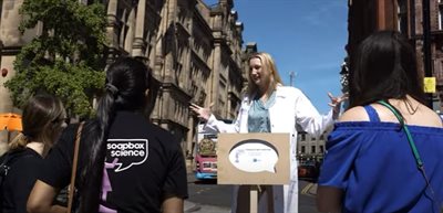Female wearing a white lab coat speaking outside to a small crowd of people
