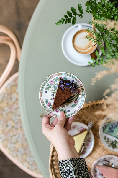 Overhead view of arm holding slice of cake on a plate and cups and saucers on table beneath