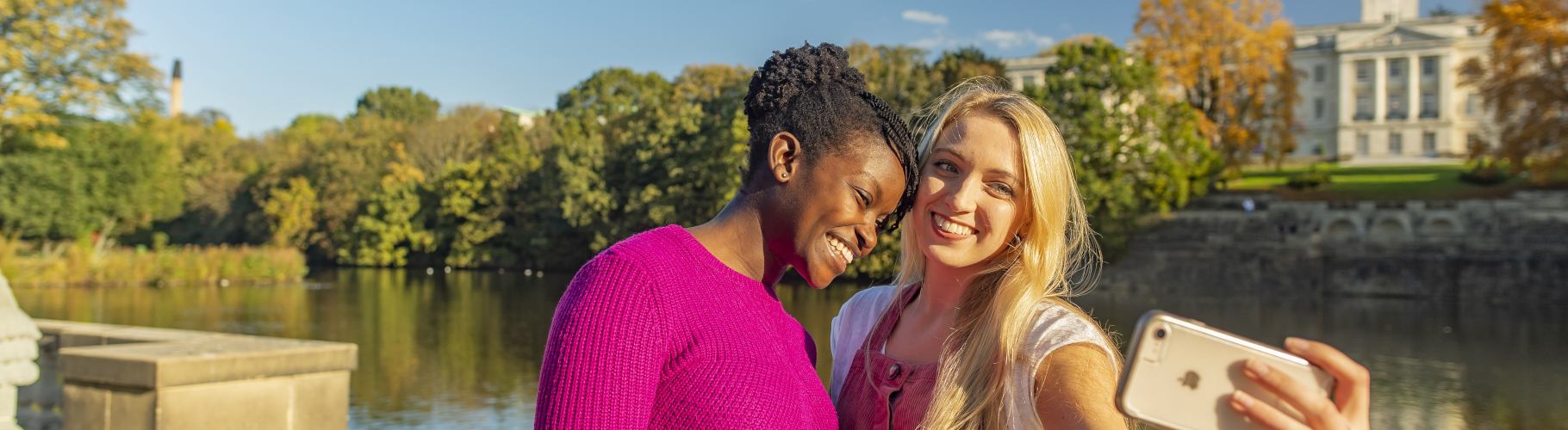 Two students take selfie in front of boating lake and Trent Building