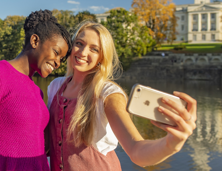 Two students take selfie in front of boating lake and Trent Building 